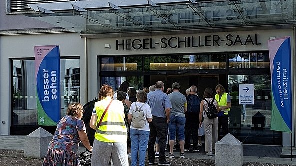 Eingangsbereich eines Veranstaltungsgebäudes mit einer Menschenmenge davor. Über dem Eingang hängt ein großes rechteckiges Plakat mit grünem Hintergrund und einer blauen, leicht ovalen Fläche in der Mitte. Darauf steht in großen weißen Buchstaben: „besser sehen“. Darunter in kleinerer Schrift: „Die Fachmesse rund ums Sehen. 3. Juli 2026 von 10 – 17 Uhr“. In der rechten oberen Ecke des Plakats befindet sich ein gelber Kreis mit der Aufschrift „Eintritt frei!“. Direkt über der Tür steht in Metallbuchstaben „Hegel-Schillersaal“. Links und rechts neben dem Eingang stehen schmale, hohe Fahnenbanner, jeweils mit blauem Feld und dem Text „besser sehen“, darunter ein kleinerer grüner Bereich. Vor dem Eingang wartet eine Schlange von Menschen auf dem gepflasterten Platz. Mehrere Personen tragen gelbe Warnwesten, andere Freizeitkleidung. Im Vordergrund schiebt eine Frau im bunten Kleid einen Rollator; neben ihr steht eine Person mit gelber Warnweste und heller Hose.