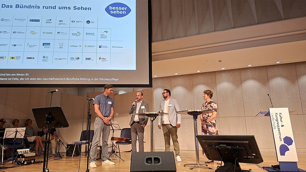 Bühnenaufnahme in einem Saal mit mehreren Personen und Präsentationsfolie. Auf einer Holz-Bühne stehen vier Personen in Freizeitkleidung an Mikrofonständern; zwei tragen helle Hosen und T-Shirts, eine Person ein bunt gemustertes Oberteil, eine weitere ein blaues T-Shirt. Links vorne stehen große Lautsprecherboxen, Mikrofonstative, ein kleineres Keyboard und weitere Technik. Rechts auf der Leinwand ist eine Präsentationsfolie projiziert. Oben steht groß: „Das Bündnis rund ums Sehen“. Darunter sind viele kleine Logos in einem Raster angeordnet. Rechts unten auf der Folie befindet sich ein rundes blaues Logo mit dem weißen Text „besser sehen“. Die Personen auf der Bühne sind v.l. Henning Müller, Felix Neher, Michael Diboun, Dr. Alexandra Sußmann.