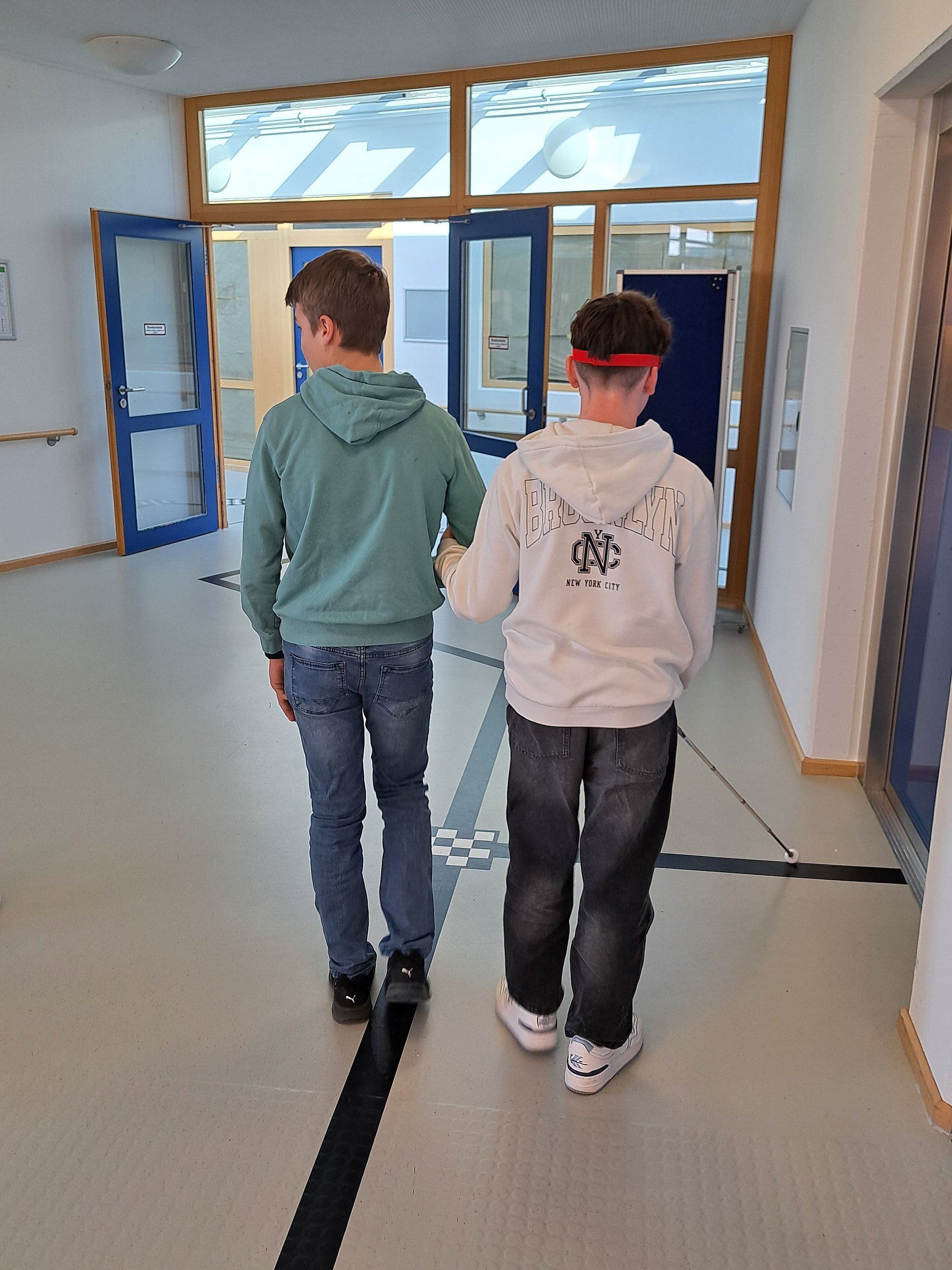Zwei Schüler laufen nebeneinander einen Flur entlang. Einer von ihnen hat seine Augen verbunden und läuft mit einem Langstock an einer Leitlinie entlang. Er hat sich mit dem Arm bei dem anderen Schüler eingehakt.