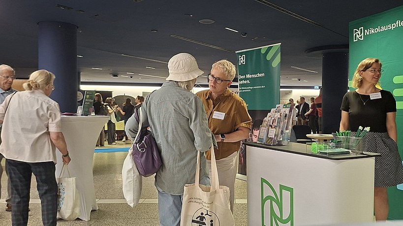 Messestand der Nikolauspflege: Im Vordergrund steht ein heller, rechteckiger Tresen mit einem grünen N-Logo auf der Front. Auf dem Tresen liegen einige flache Gegenstände und mehrere kleine Give-aways. Davor stehen unten im Bild zwei Personen an der Thekenseite: links eine Person in dunkelblauem Oberteil und dunklem Rock, rechts eine Person in dunkler Kleidung, beide mit Namensschildern auf der Brust. Hinter ihnen bildet eine große, grüne Rückwand mit weißem Logo und Schrift den Hintergrund des Standes. Auf der Wand ist der Schriftzug „Nikolauspflege“ zu erkennen. In der Bildmitte stehen zwei ältere Personen, eine mit hellgrauer Jacke und hellen Haaren, die nach hinten gebunden sind, eine andere mit braunem Hemd und grauen Haaren. Beide tragen Namensschilder und wirken so, als würden sie sich mit den Standpersonen unterhalten. Eine Person hat eine Stofftasche mit einem dunklen Motiv über der Schulter, eine andere eine lila Umhängetasche.