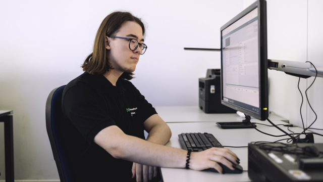 Ein junger Mann in einem schwarzen T-Shirt sitzt an einem Schreibtisch. Er hat schulterlange braune Haare und trägt eine Brille. Mit seiner rechten Hand bedient er eine Maus. Er blickt auf einen Computerbildschirm