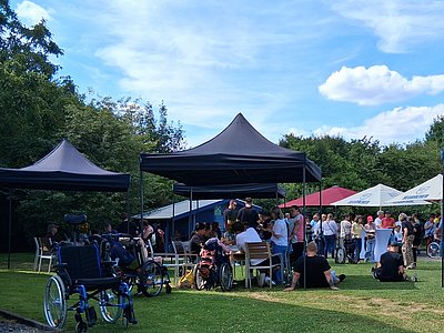 Begegnung am Limeshof. Verschiedene bunte Pavillon und Menschen in entspannter Atmosphäre. Der Himmel ist blau, viel grüne Baume sind zu sehen und ein paar weiße Wölkchen.