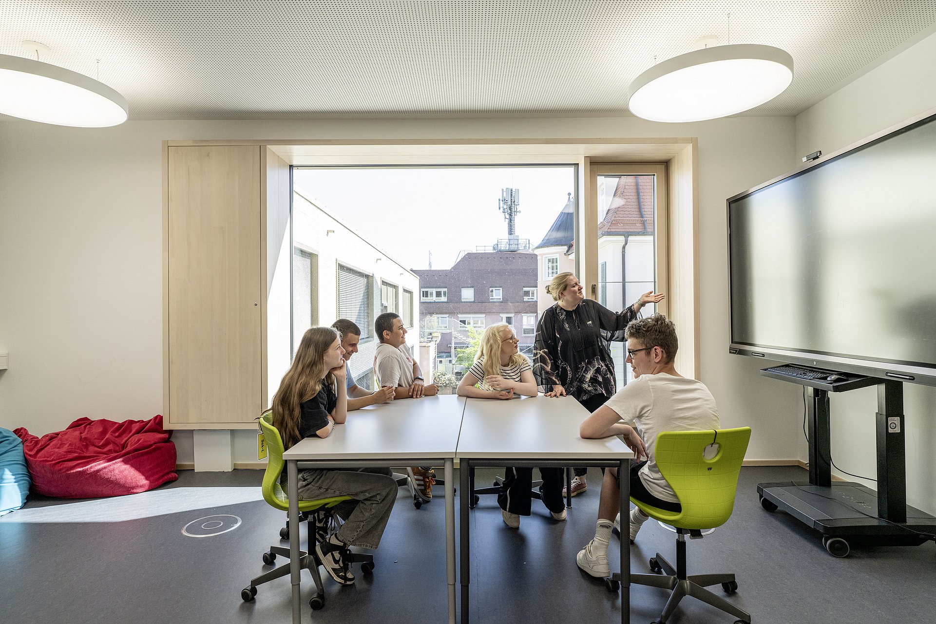 Eine Gruppe von Jugendlichen sitzt in einem Klassenzimmer zusammen an einem Tisch. Eine Lehrerin erklärt gerade etwas am Smartboard und alle hören aktiv zu.