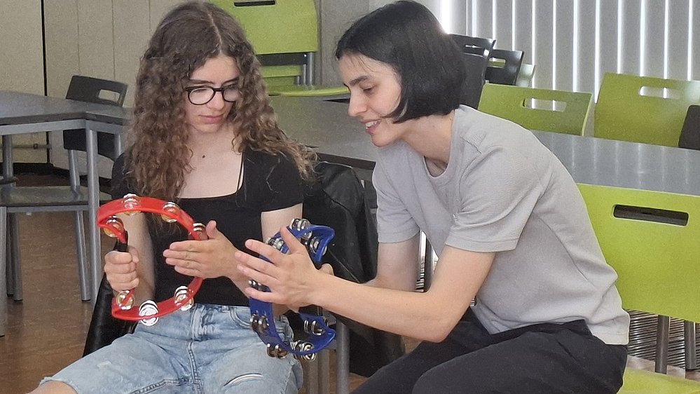 Zwei sitzende Personen mit Tamburins. Links sitzt eine junge Frau mit langen braunen Locken und schwarzer Brille. Sie trägt ein schwarzes Oberteil und eine hellblaue Jeans mit großen Rissen an beiden Knien. In den Händen hält sie ein rotes Tamburin mit Metallschellen, das sie etwas schräg nach vorn hält. Rechts daneben sitzt eine zweite Frau mit kurzem, dunklem Haar. Sie trägt ein hellgraues T‑Shirt und eine dunkle Hose. Diese Person beugt sich leicht nach links zur ersten Person hin, lächelt und hält ein blaues Tamburin, das ein Stück näher an die andere Person herangeführt wird. Beide sitzen auf einfachen Hockern in einem Raum mit Holzfußboden und Tischen und Stühlen im Hintergrund.