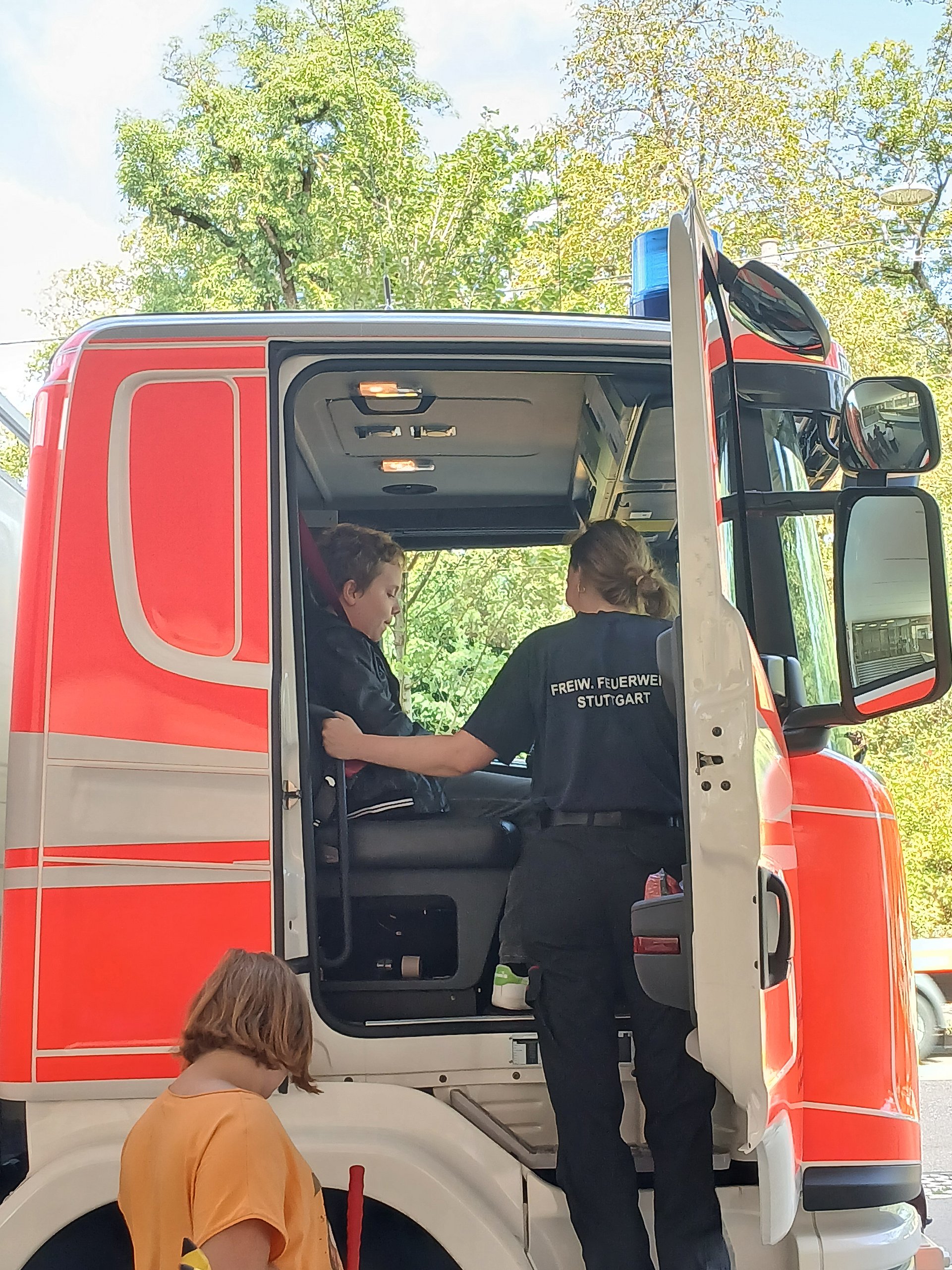 Ein Feuerwehrauto steht draußen mit geöffneter Tür. Eine Frau steht in der Tür. Im Innenraum des Autos sitzt ein Schüler auf dem Beifahrersitz