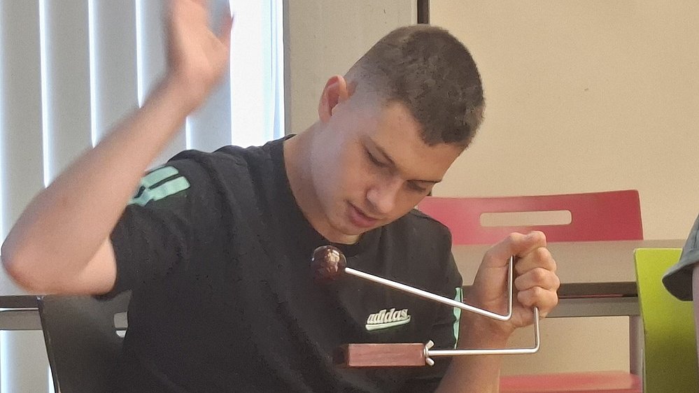 Ein junger Mann sitzt auf einem Stuhl und bedient eine kleine Triangel. Er trägt ein schwarzes T Shirt mit einem weißen „adidas“-Logo auf der Brust und hat kurz geschnittene, dunkle Haare. Sein Kopf ist leicht nach vorn und unten geneigt, der Blick geht konzentriert auf das Instrument in seiner rechten Hand. Das Instrument besteht unten aus einem flachen rechteckigen Holzblock, aus dem ein gebogener Metallbügel nach oben führt. Oben am Metallbügel ist eine dunkle Kugel befestigt, die wie eine kleine Holzkugel wirkt. Die linke Hand des Mannes ist angehoben, als würde er gleich auf die Kugel oder den Bügel schlagen oder eine Bewegung ausführen.   Im Hintergrund stehen einfache Stühle, einer mit roter Rückenlehne.