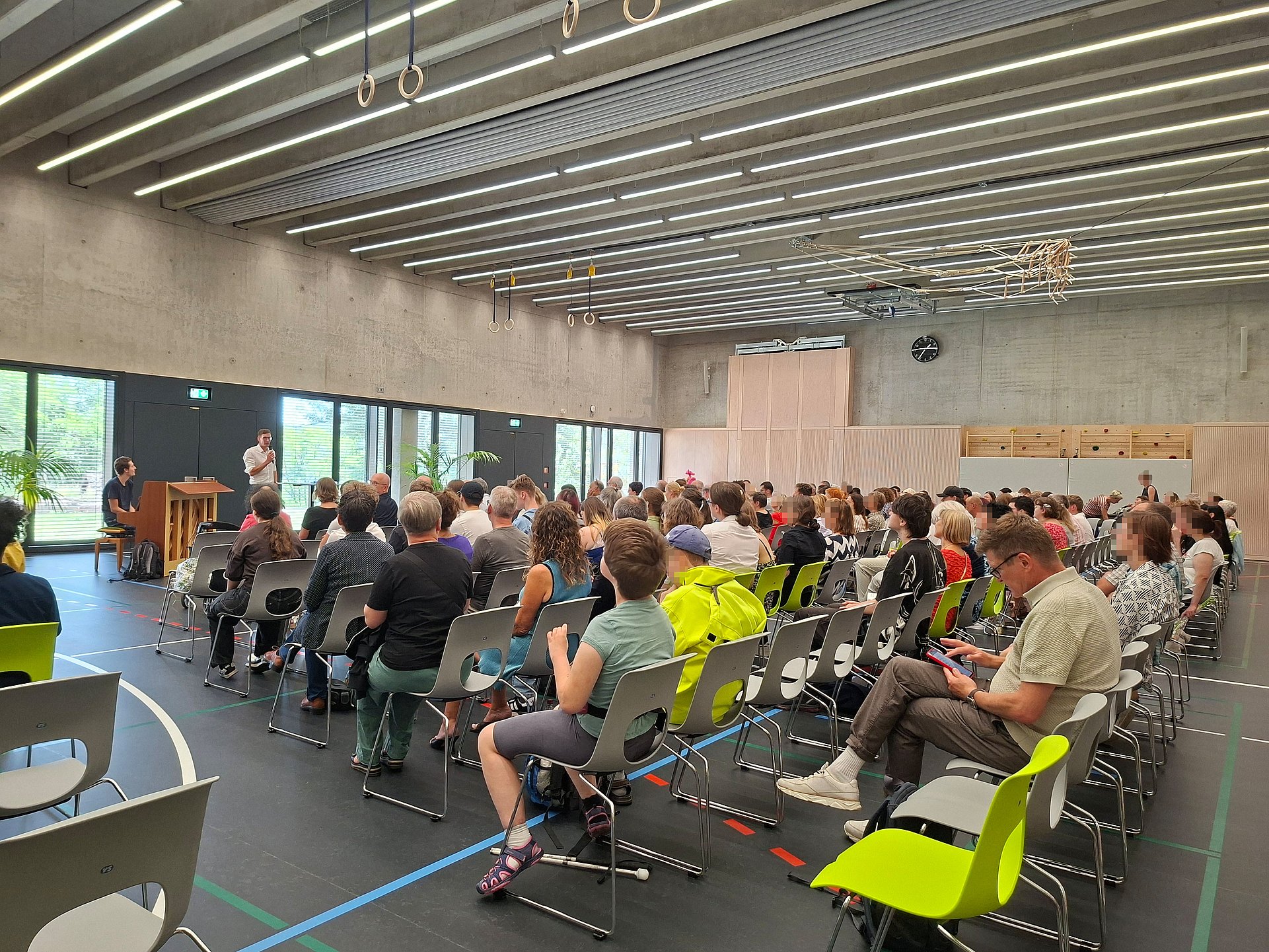 Viele Menschen sitzen in Stuhlreihen hintereinander in der Sporthalle. Vor ihnen sitzt ein Mann an einem Klavier. Daneben spricht der Schulleiter in ein Mikrofon