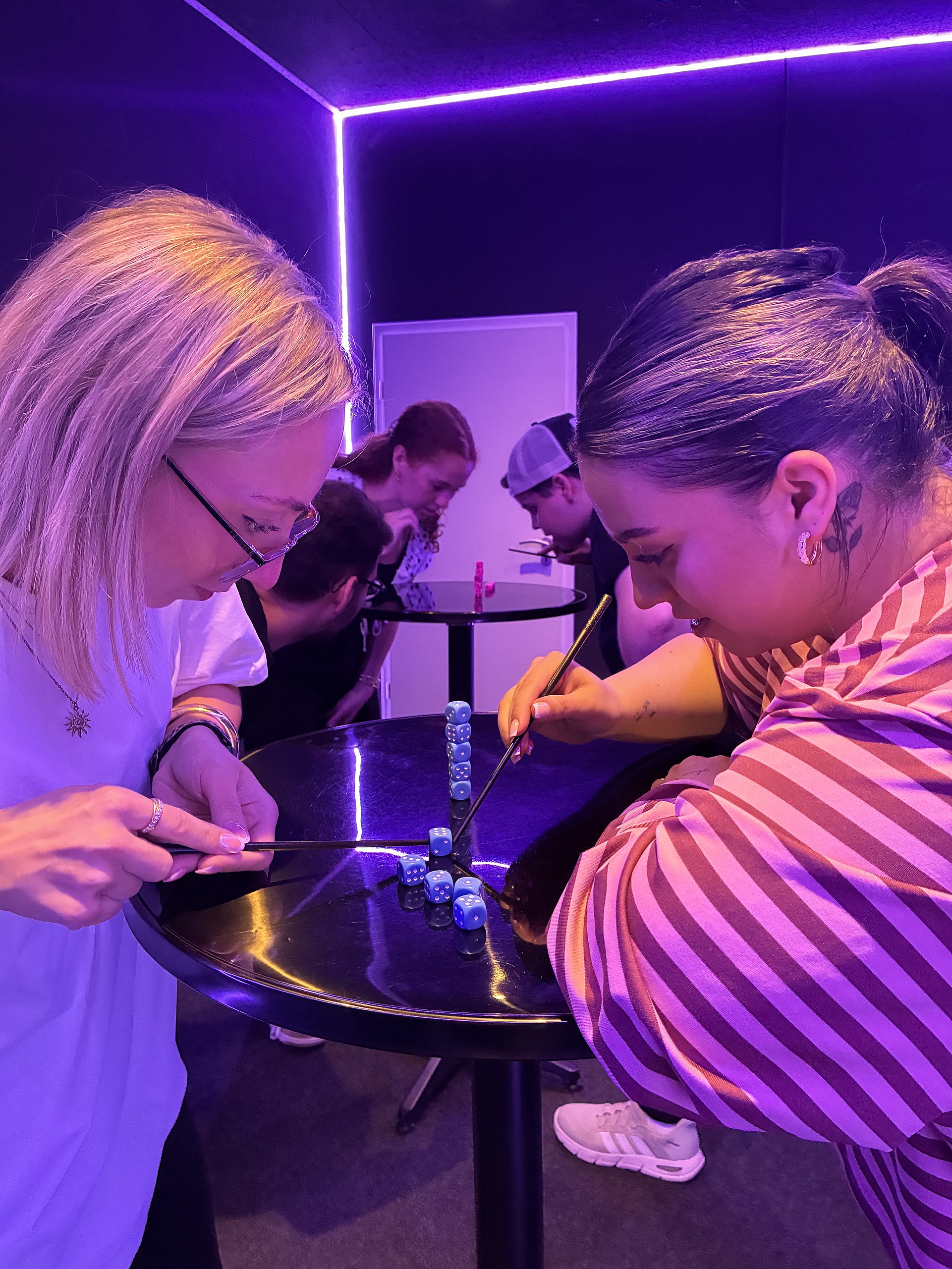 Zwei Frauen an einem hohen, runden, schwarzen Tisch in einem Raum mit violetter Neonbeleuchtung. Auf der Tischplatte liegen mehrere kleine, blau‑weiße Würfel mit Punkten, manche stehen übereinandergestapelt zu einem kleinen Turm. 