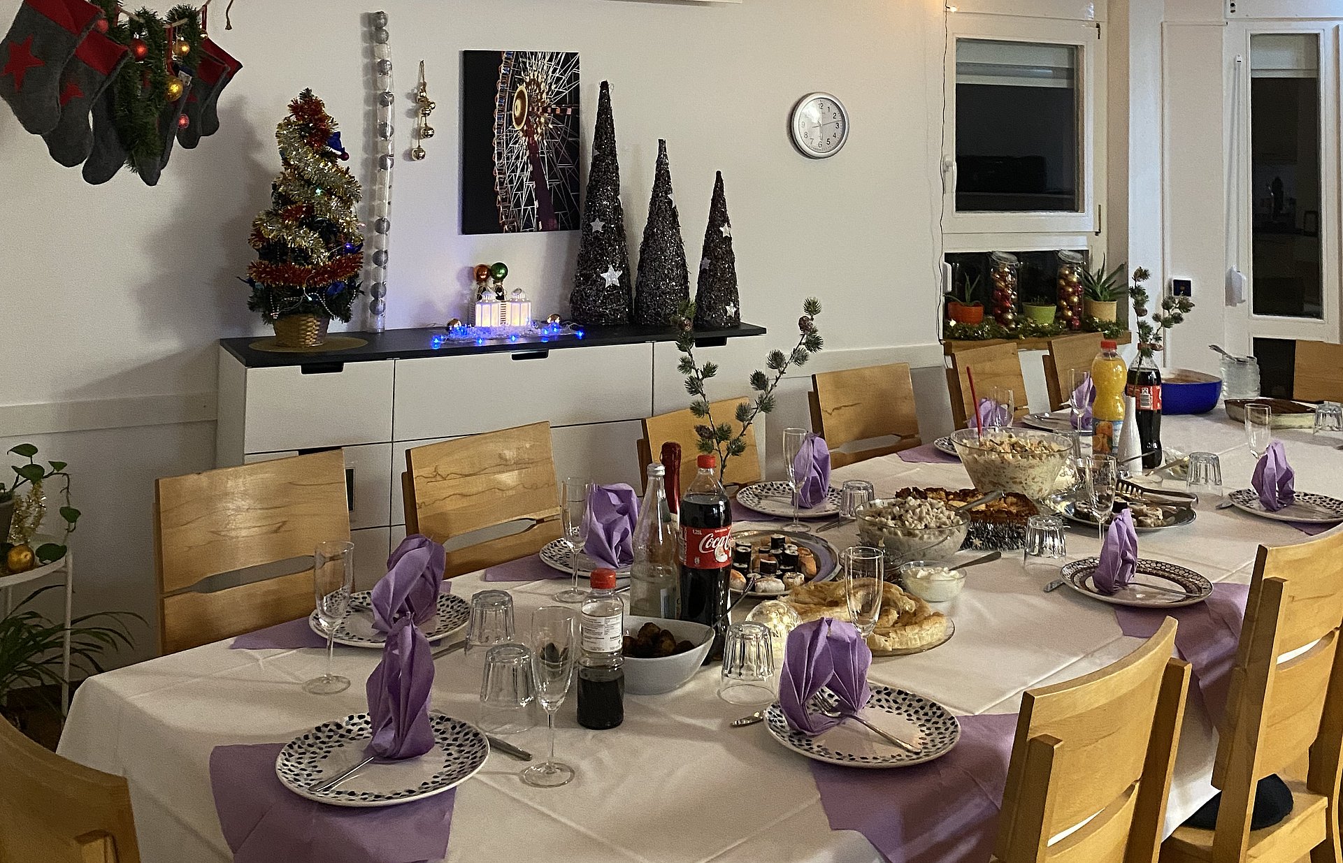 Ein gedeckter Tisch mit einer weißen Tischdecke steht in einem Zimmer. Darauf stehen Gläser, Teller, Besteck, Servietten, Blumen und Kerzen. 