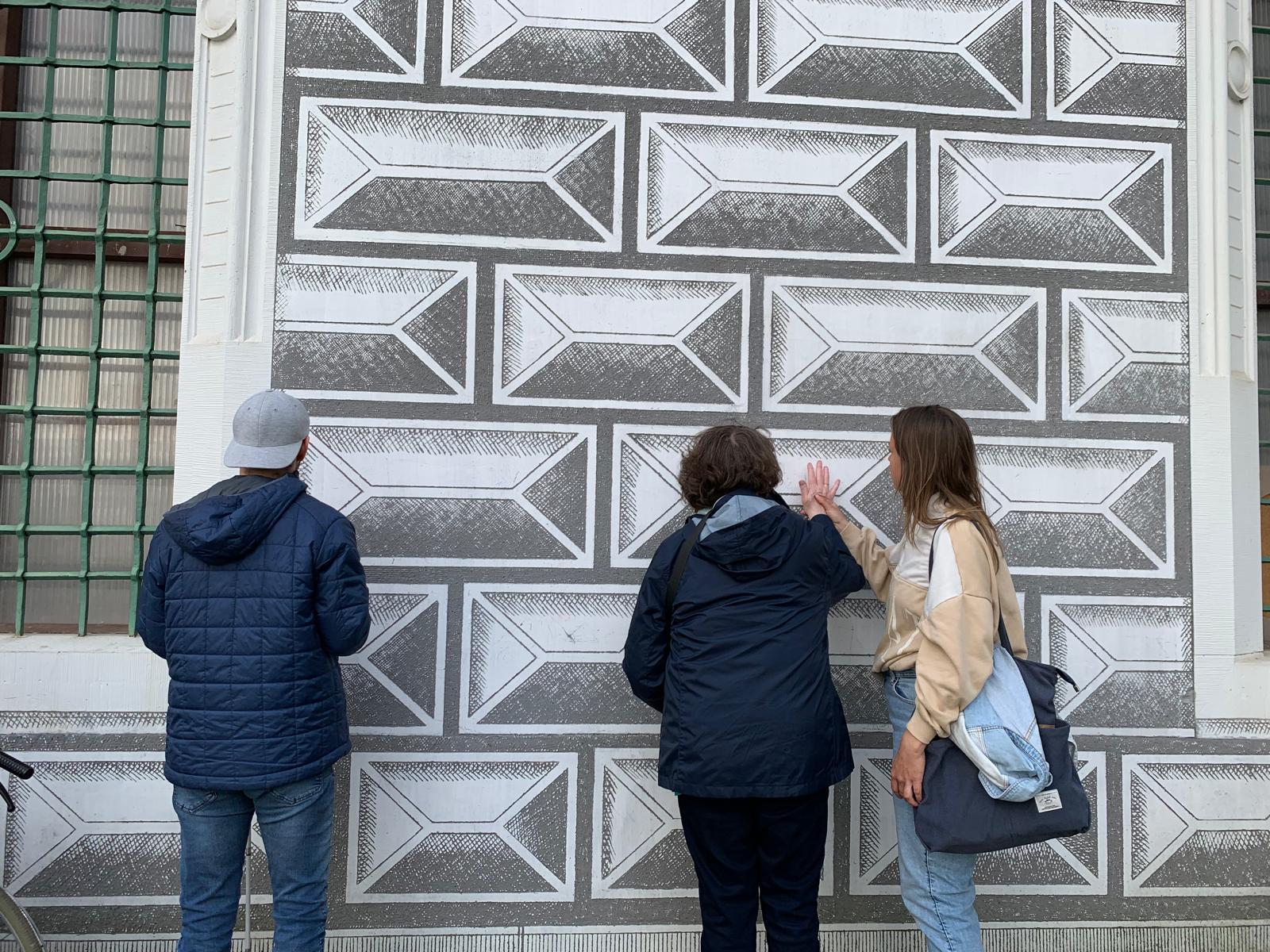 Zwei Personen stehen vor einer Wand mit grafischem Muster. Die Wand ist mit vielen rechteckigen Feldern dekoriert, jedes Rechteck hat innen eine Art schräg gezeichnetes Rahmenmotiv, sodass ein 3D‑Effekt entsteht. Die Flächen sind in Grautönen gehalten, mit schraffierten Bereichen, die wie Steinquader wirken. Links steht eine Person mit mittelkurzen, dunklen, leicht lockigen Haaren. Diese Person trägt eine dunkle, navyblaue Jacke und eine schwarze Hose.